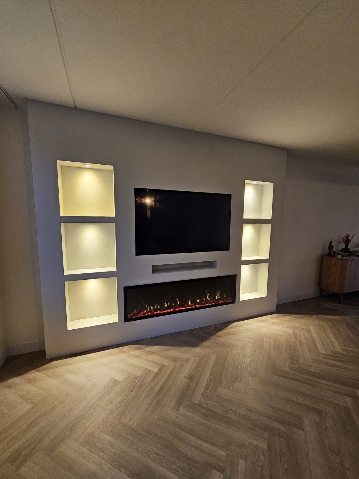 Witte cinewall met sfeerhaard en verlichte nissen in Almere 3 Cinewall in Almere schuin van links met brandende sfeerhaard en verlichte nissen
